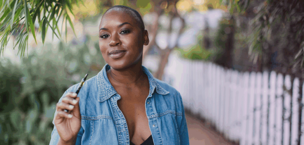 Confident woman outdoors holding a cannabis vape pen, styled casually in a denim shirt.