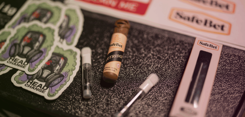Close-up of cannabis vape products, cartridges, and branded stickers from SafeBet and Head Change on a textured surface.
