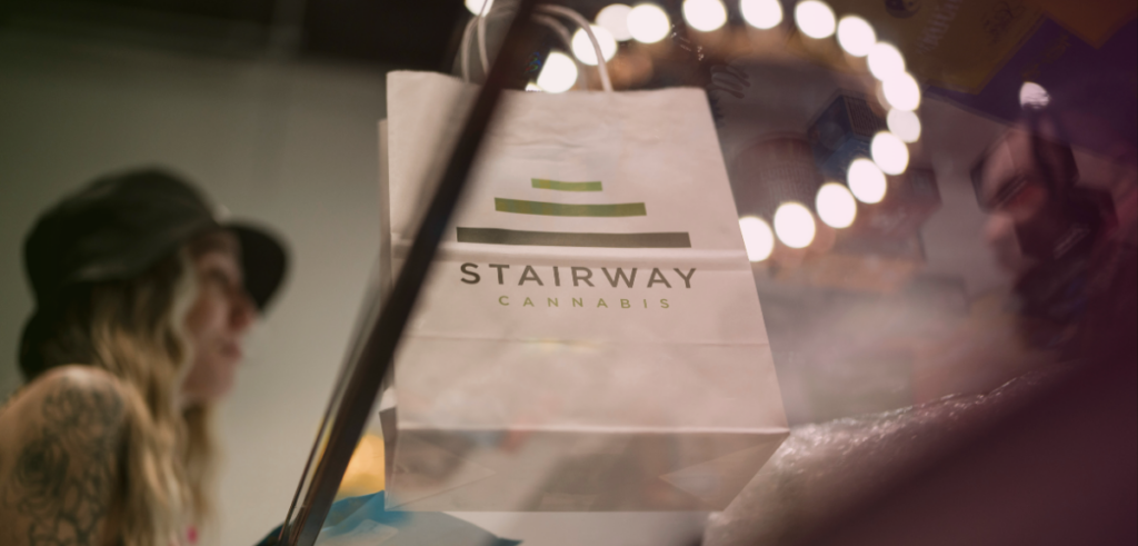 A low-angle shot of Kate, a budtender at Stairway Cannabis, standing behind the sales counter with a bag already placed on the counter, ready for a customer.