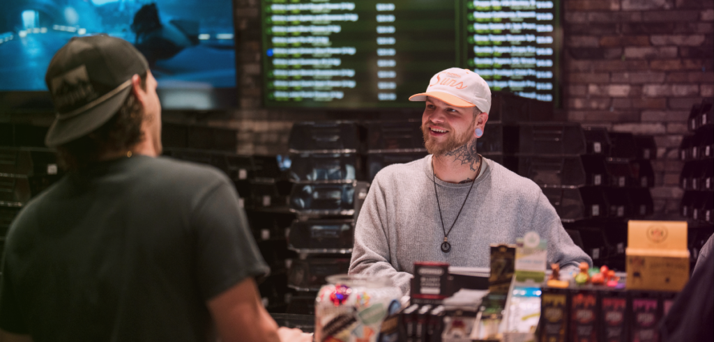 Mo, a budtender at Stairway Cannabis, stands behind the sales counter, smiling while conversing with a recreational customer about cannabis.