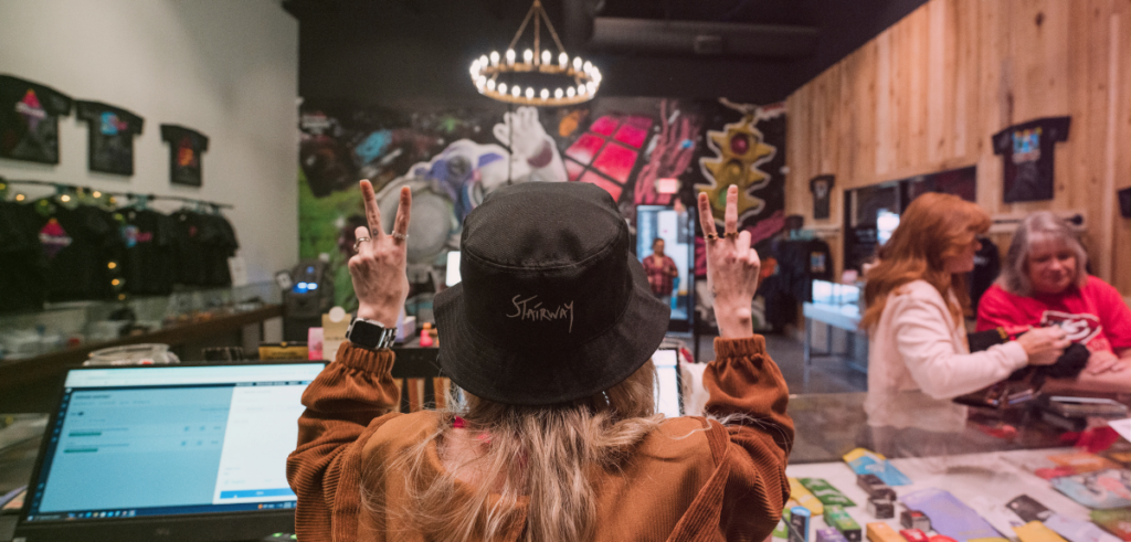 A female budtender at Stairway Cannabis stands behind the sales counter, holding up two peace signs while looking out at the sales floor.