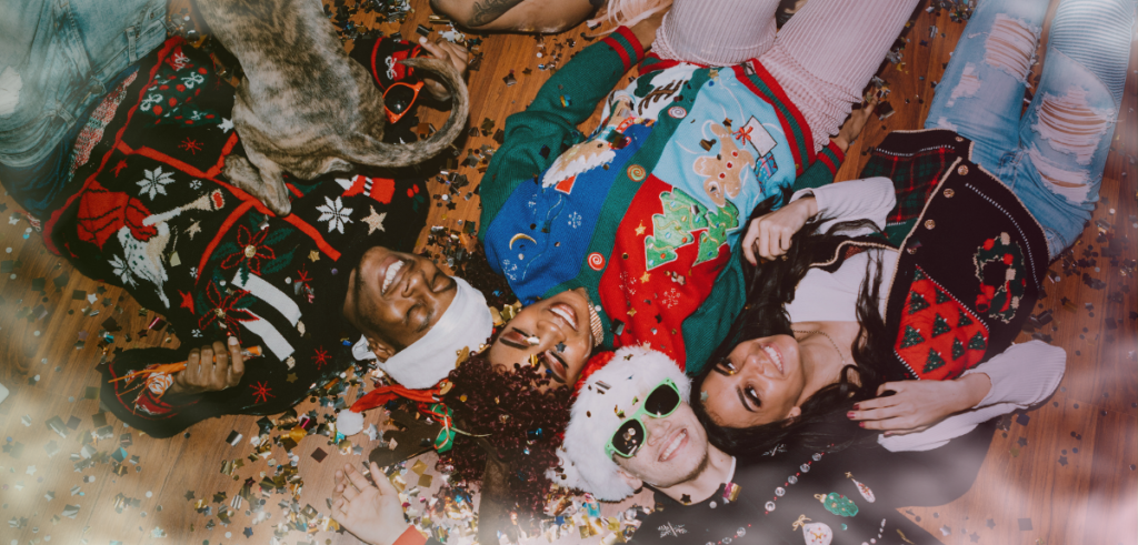 An overhead view of friends wearing festive sweaters and a Santa hat while lying on the floor during a holiday party.