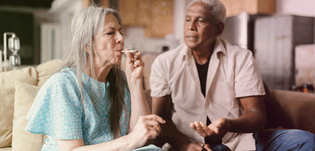 Two people sitting on a couch, one smoking a pre-roll and one looking on at her.