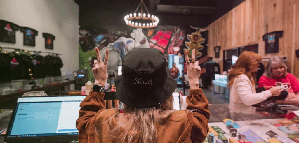 A female Stairway Cannabis budtender stands behind the sales counter looking out at the sales floor while holding up two peace signs.