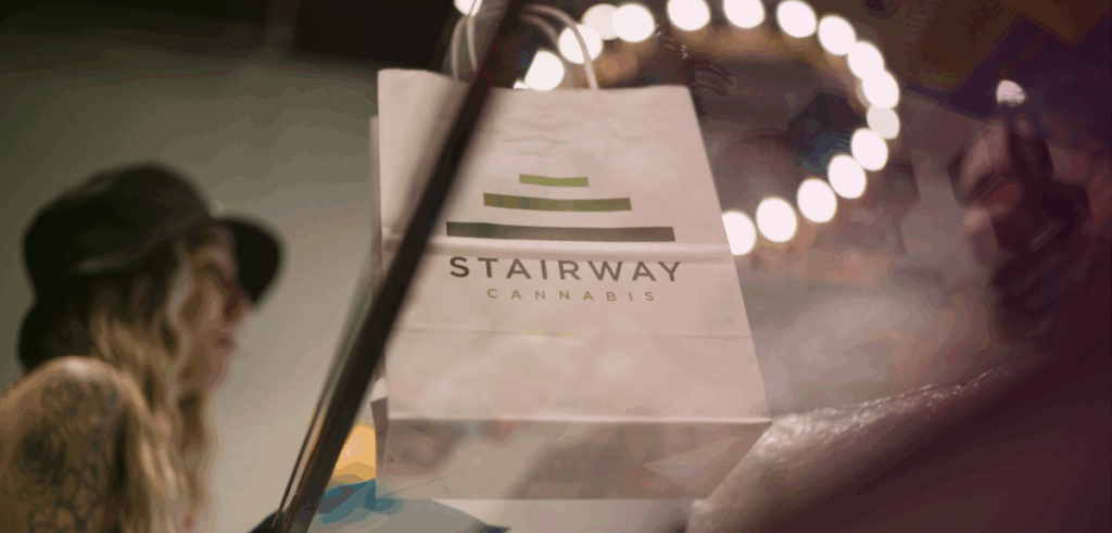 A low-angle shot of Kate, a budtender at Stairway Cannabis, standing behind the sales counter with a bag already placed on the counter, ready for a customer.