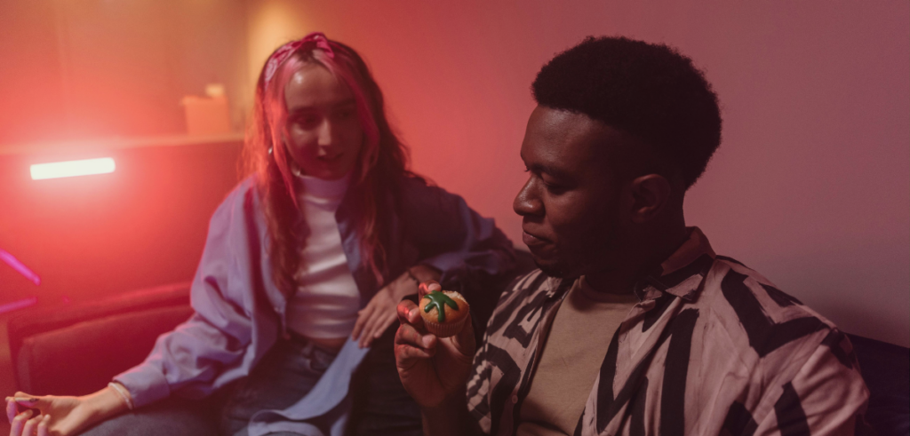 A black man holds a cannabis-themed muffin with a green marijuana leaf decoration while sitting in a dimly lit, red-hued setting, casually chatting with a woman.