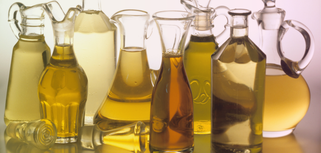 Assorted glass bottles filled with different types of cooking oils, varying in color from light yellow to deep amber, arranged against a neutral background.