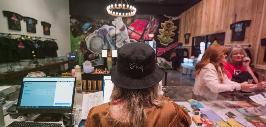 A female Stairway Cannabis budtender stands behind the sales counter looking out at the sales floor.