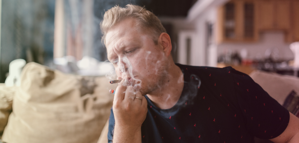 A close up of a white male smoking a joint with smoke billowing in his face.