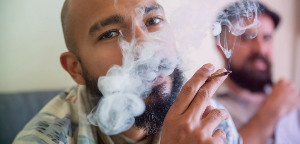 Close-up of a Black man exhaling a cloud of smoke while holding a joint, with a blurred white man in the background.