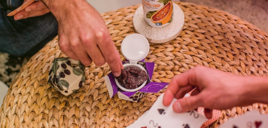 The above view point of a male hand reaching into a container of Wyld edibles while a second hand plays with a deck of cards.