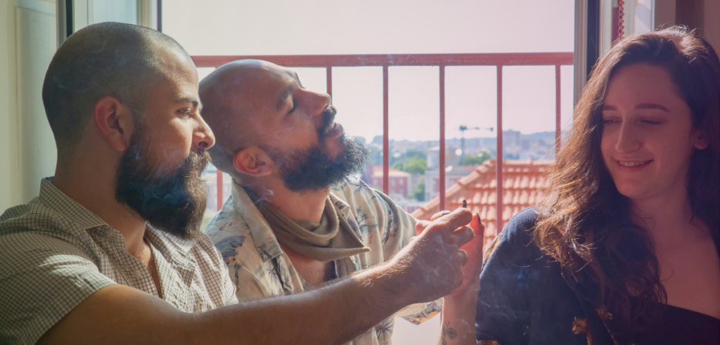 3 people on a balcony passing a lit pre-roll.