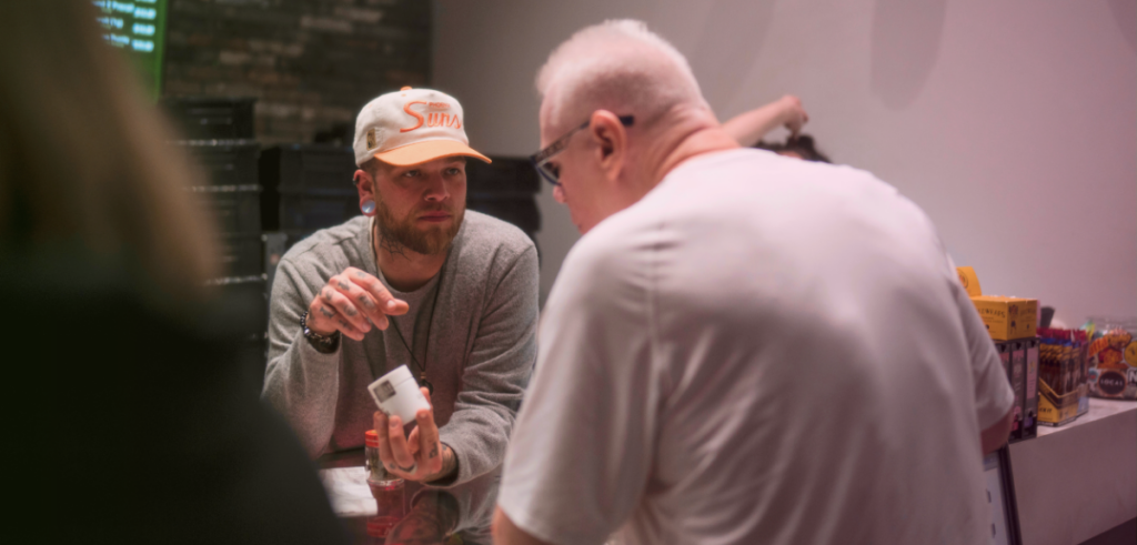 Budtender Mo holds a jar of cannabis flowers as he talks to a middle-aged male customer at the sales counter.