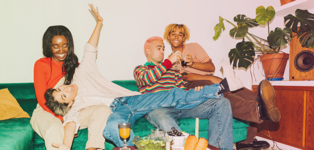 Four friends relax and laugh together on a green couch, enjoying drinks and snacks in a cozy, plant-filled space.