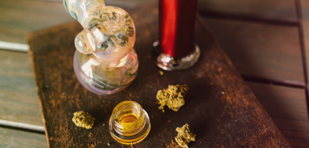 A glass pipe, cannabis buds, and a small jar of concentrate sit on a wooden surface.