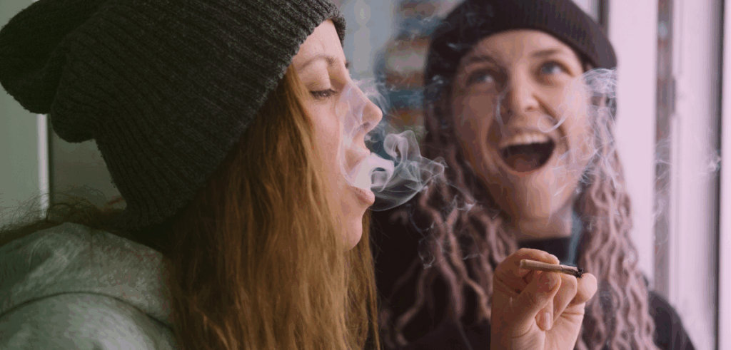 A woman blows out smoke with a female friend behind her laughing.