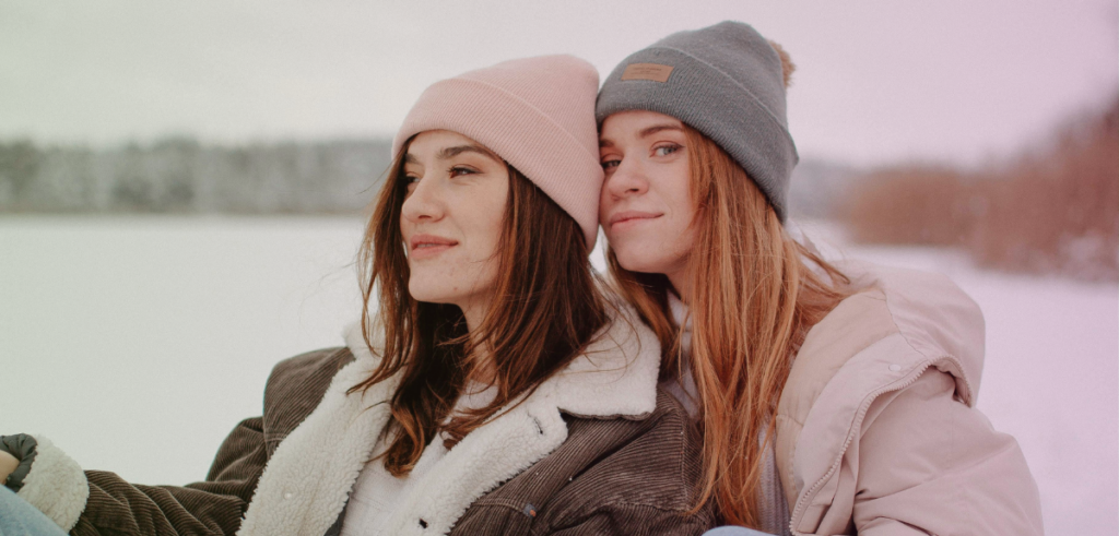 Two female-identifying people sit with stocking caps on in a winter outdoor scene.